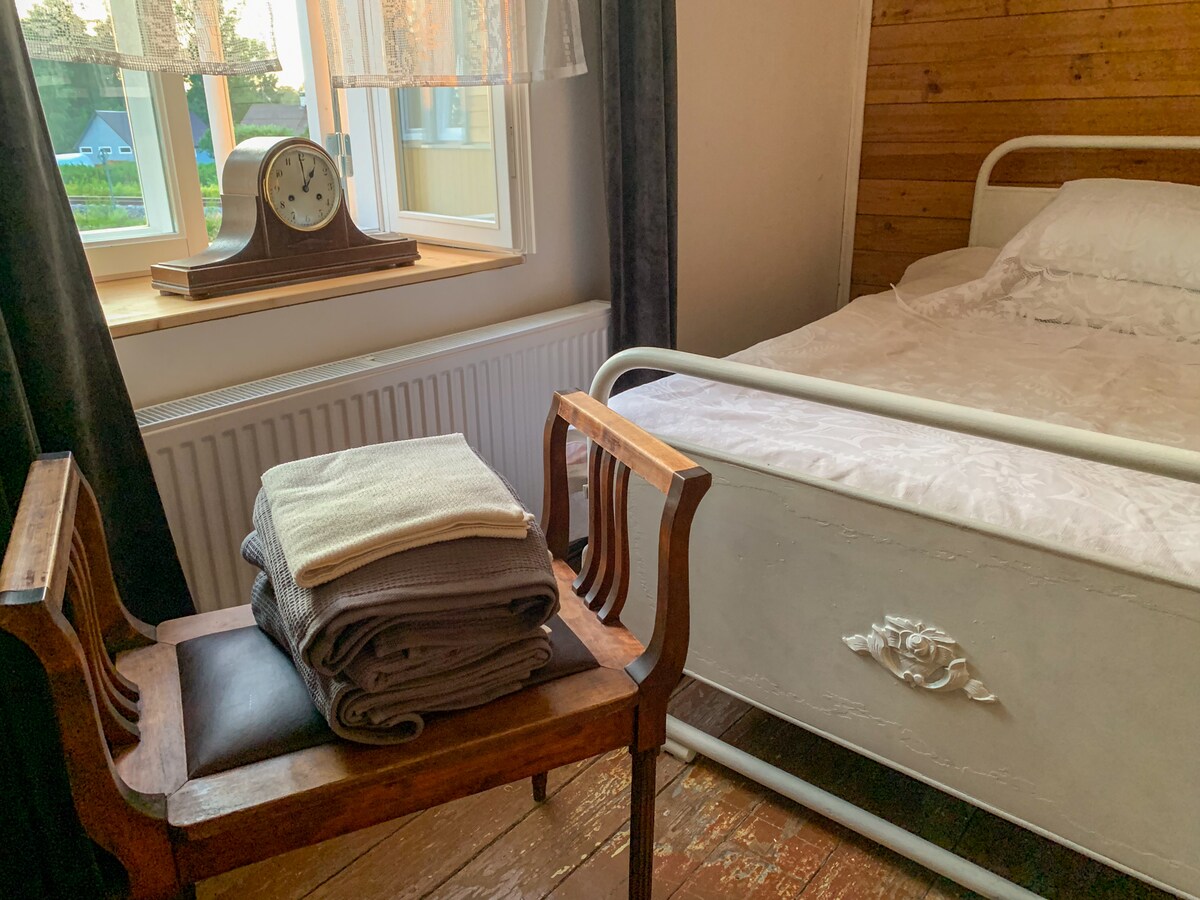 A cozy bedroom features a vintage white metal bed with soft linens. Next to it, a wooden chair holds neatly folded towels. Sunlight filters through a window adorned with light curtains, illuminating the warm wood accents of the room.