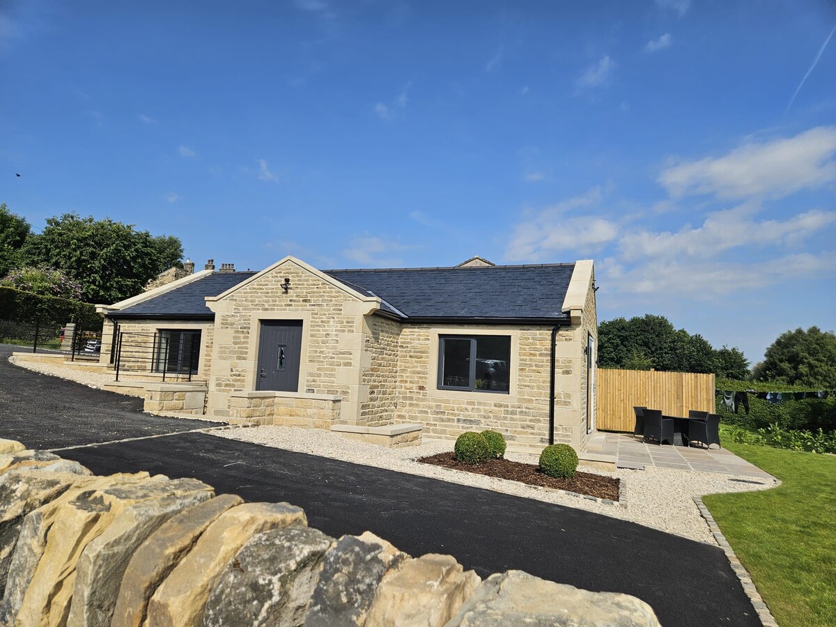 A modern holiday cottage is shown from the exterior, featuring a stone façade and a slate roof. The structure is surrounded by a gravel driveway with landscaped areas, including small shrubs. Clear blue skies and a well-maintained yard enhance the inviting appearance of the property.