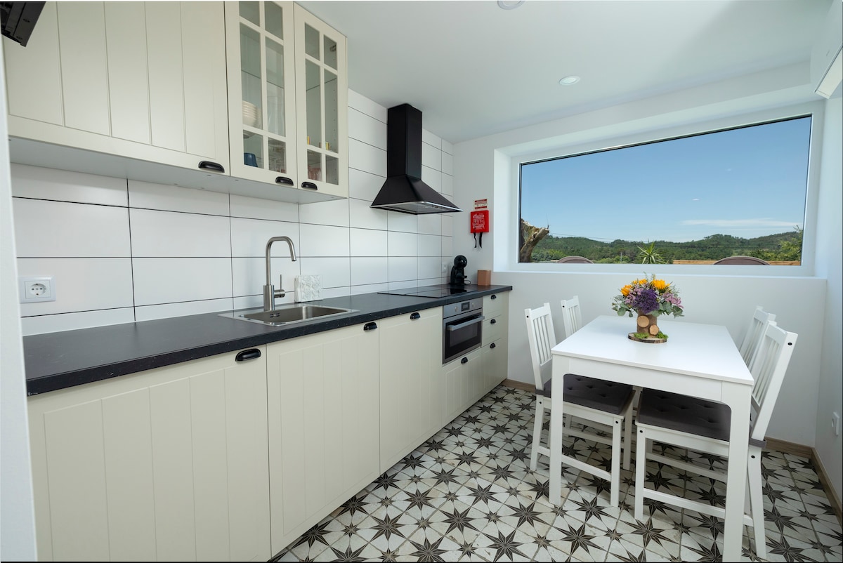A well-equipped kitchen features modern appliances, including an oven and coffee maker. Light-colored cabinetry complements the patterned floor tiles. A dining table with white chairs is positioned near a large window that offers a view of the surrounding landscape.