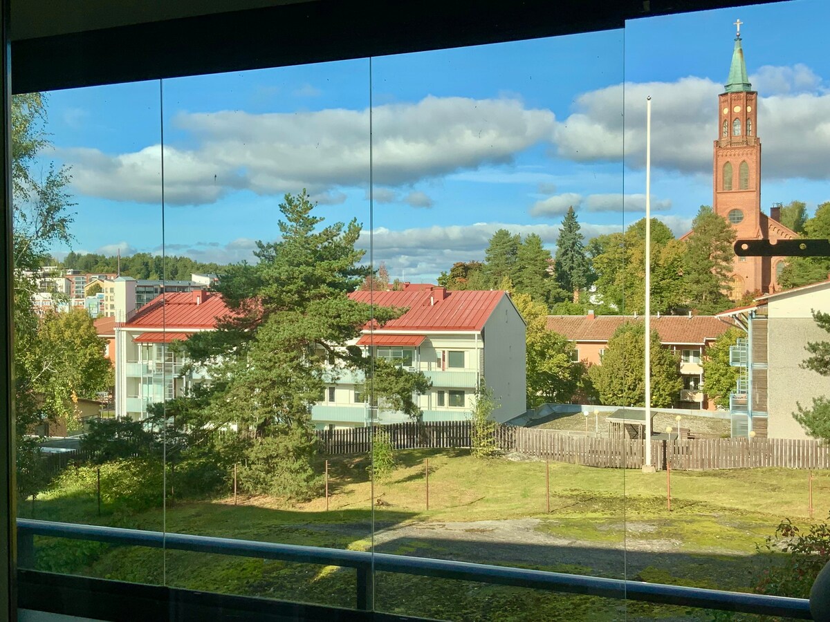 A view from the apartment showcases a green outdoor space framed by trees and a nearby building. In the background, a prominent clock tower stands above the landscape, with clear blue skies accented by soft clouds.