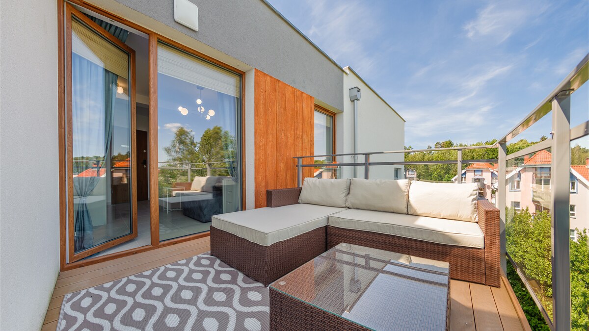 A balcony area features a sectional sofa with plush cushions and a glass-top table. The wooden flooring is complemented by a patterned rug. Large sliding doors provide access to the interior, inviting ample natural light and offering views of the surrounding greenery.