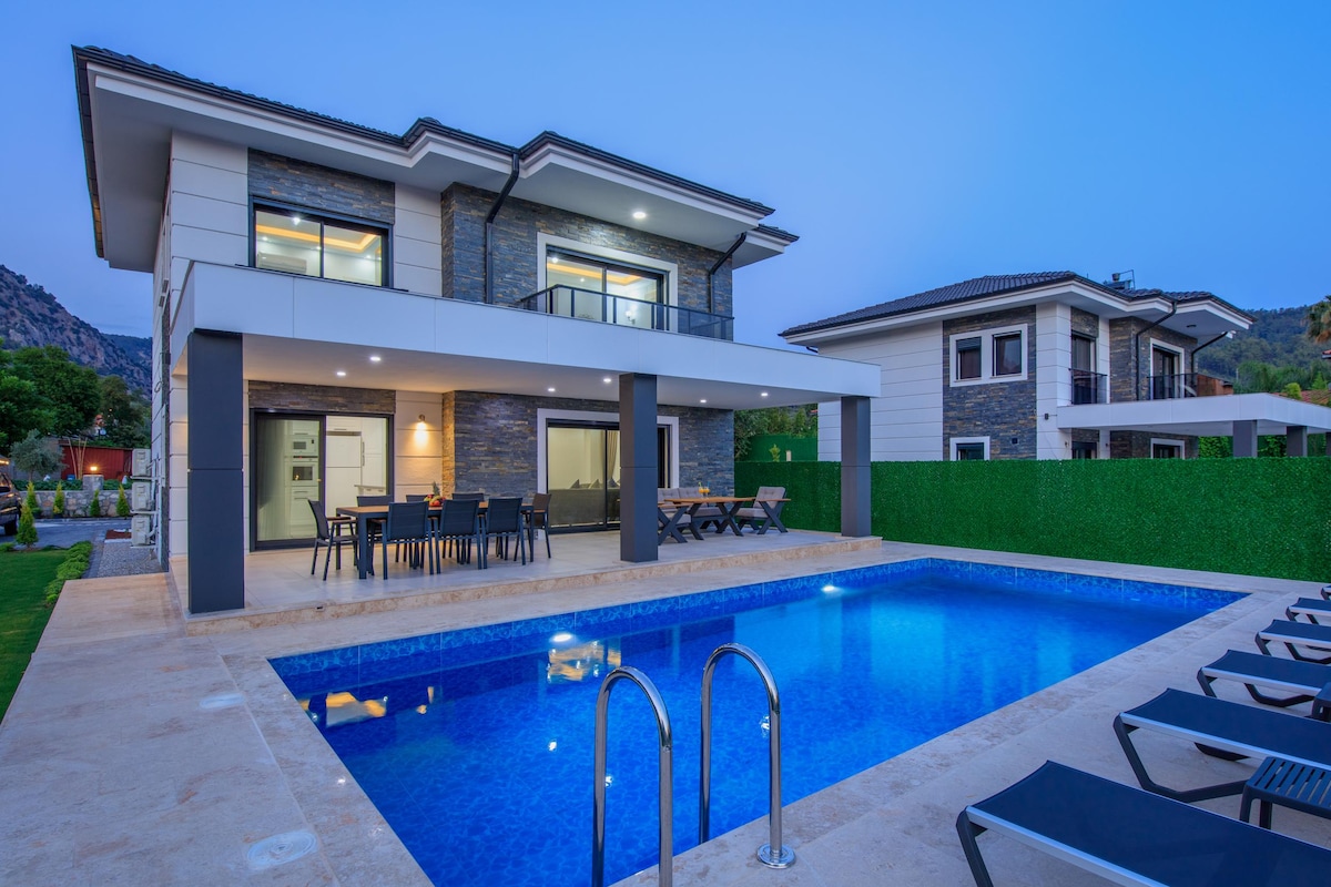 A modern villa is displayed, featuring a swimming pool with a clear blue surface. Surrounding the pool, sun loungers are arranged. The exterior of the villa highlights a blend of stone and wood materials, with large glass doors that provide access to an outdoor dining area.