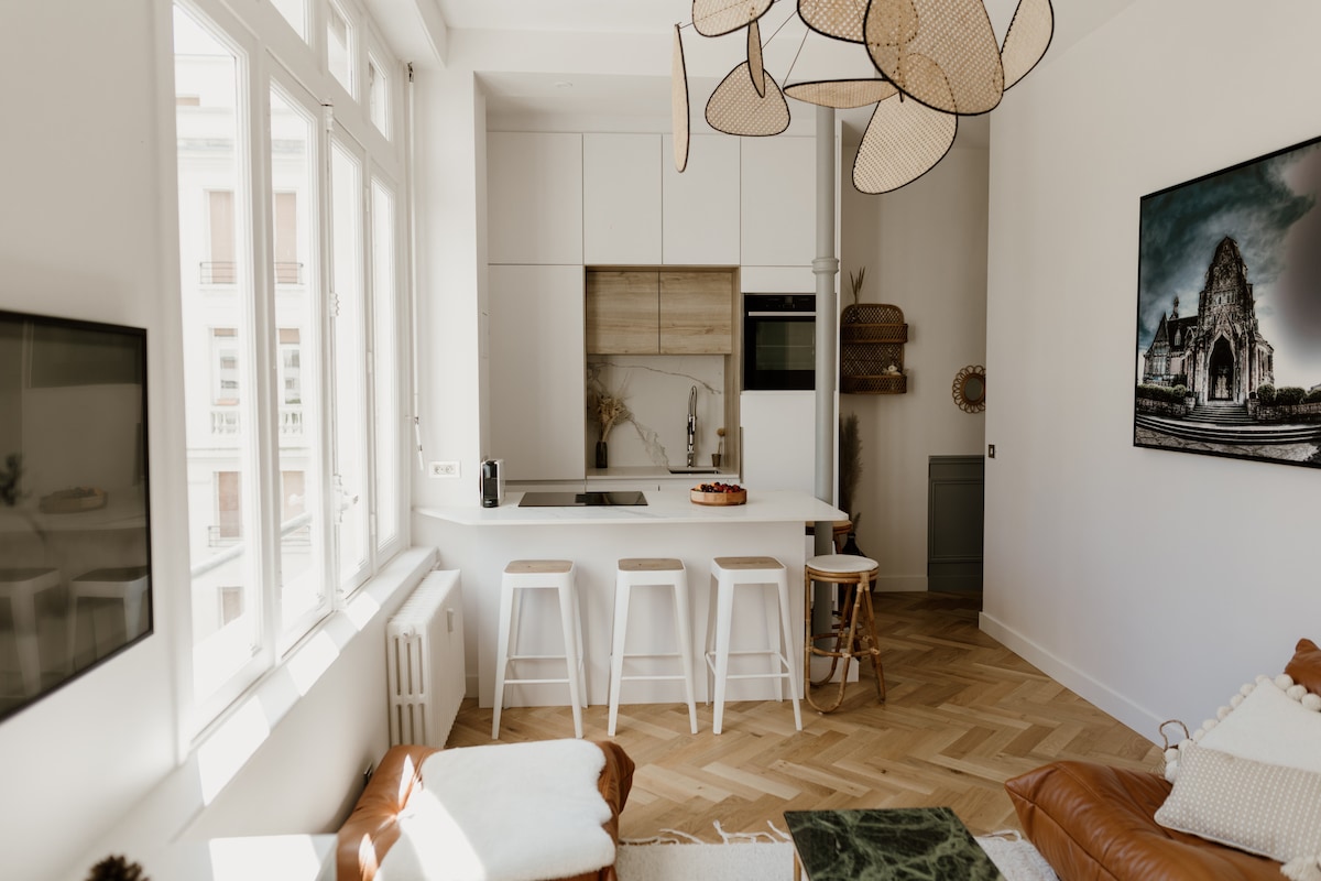 A light and airy kitchen area features modern cabinetry and an island with seating. Natural light flows in through large windows, illuminating the herringbone wooden flooring. Decorative elements, including a woven light fixture, enhance the contemporary feel of the space.