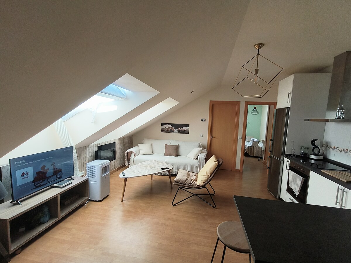 An inviting living space features a light wood floor and sloped ceiling. A cozy sofa sits beside a modern coffee table, while a television occupies a nearby shelf. Natural light filters through skylights, creating a warm ambiance throughout the area.