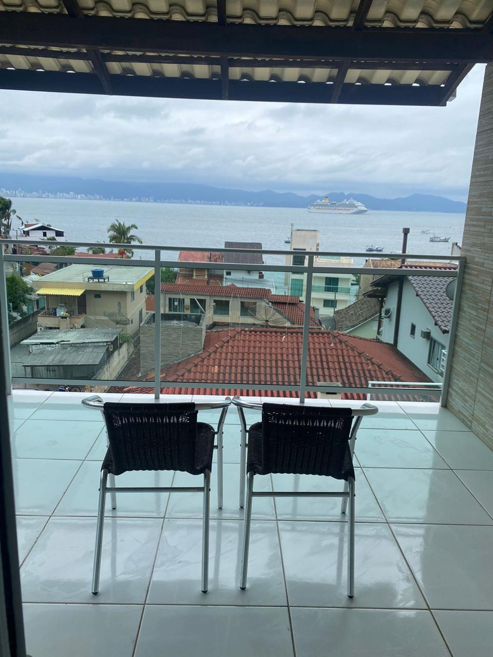 A balcony featuring two chairs is positioned to offer a view of the sea and nearby rooftops. The area is partially covered by a roof, providing shade while allowing natural light to illuminate the space. The serene coastal scenery enhances the relaxing atmosphere.