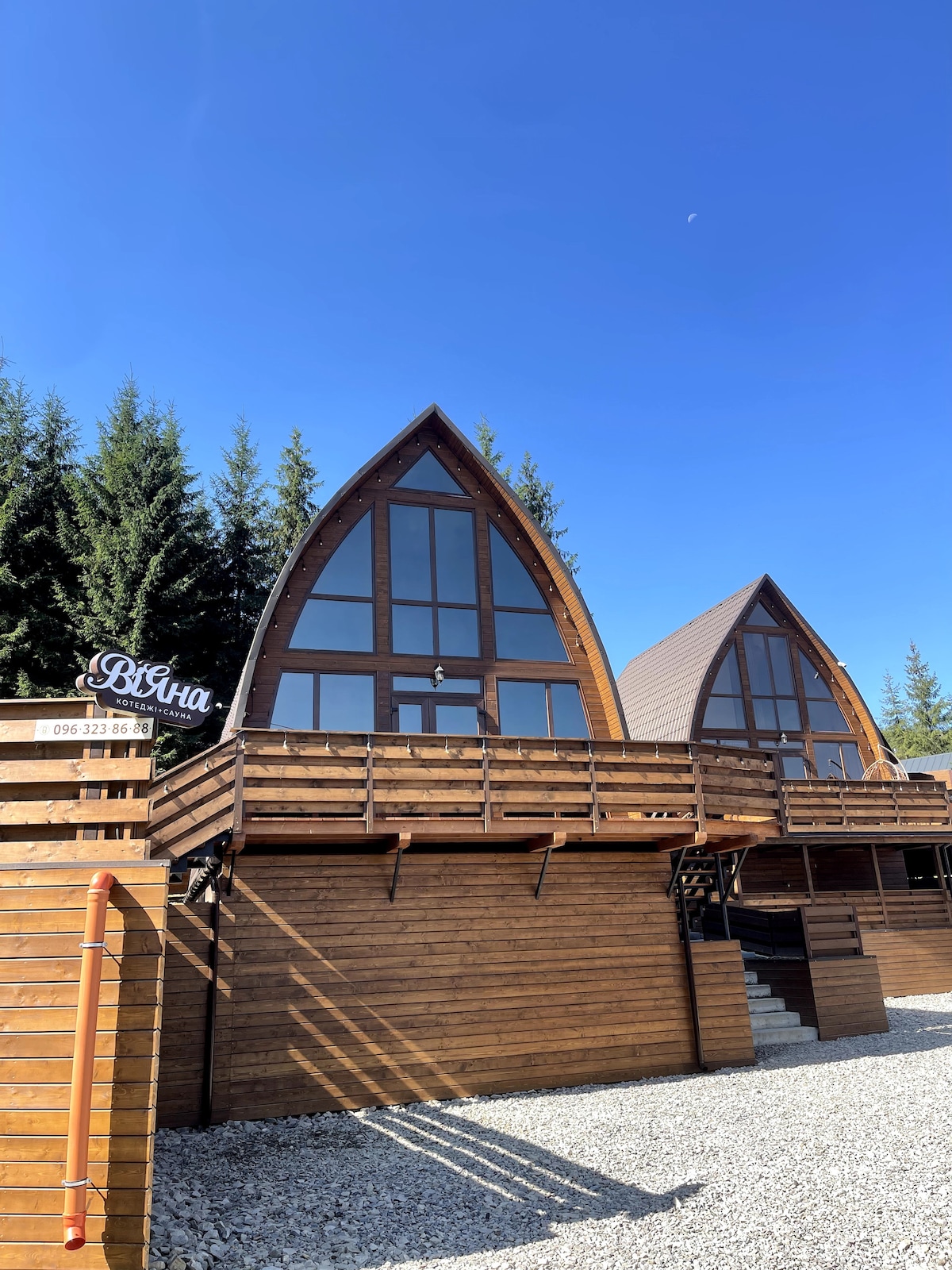 The exterior features two distinct wooden cottages with sharp, triangular roofs under a clear blue sky. Large windows allow for ample natural light. A simple wooden sign displays the name 'ВіЯна'. Surrounding evergreen trees add a natural backdrop.