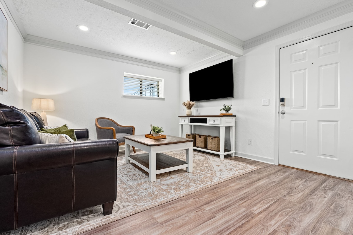 A cozy living area features a dark leather sofa paired with a wooden coffee table. A flat-screen TV is mounted on the wall, and a decorative plant is placed on the table. Natural light enters through a window, enhancing the welcoming atmosphere.