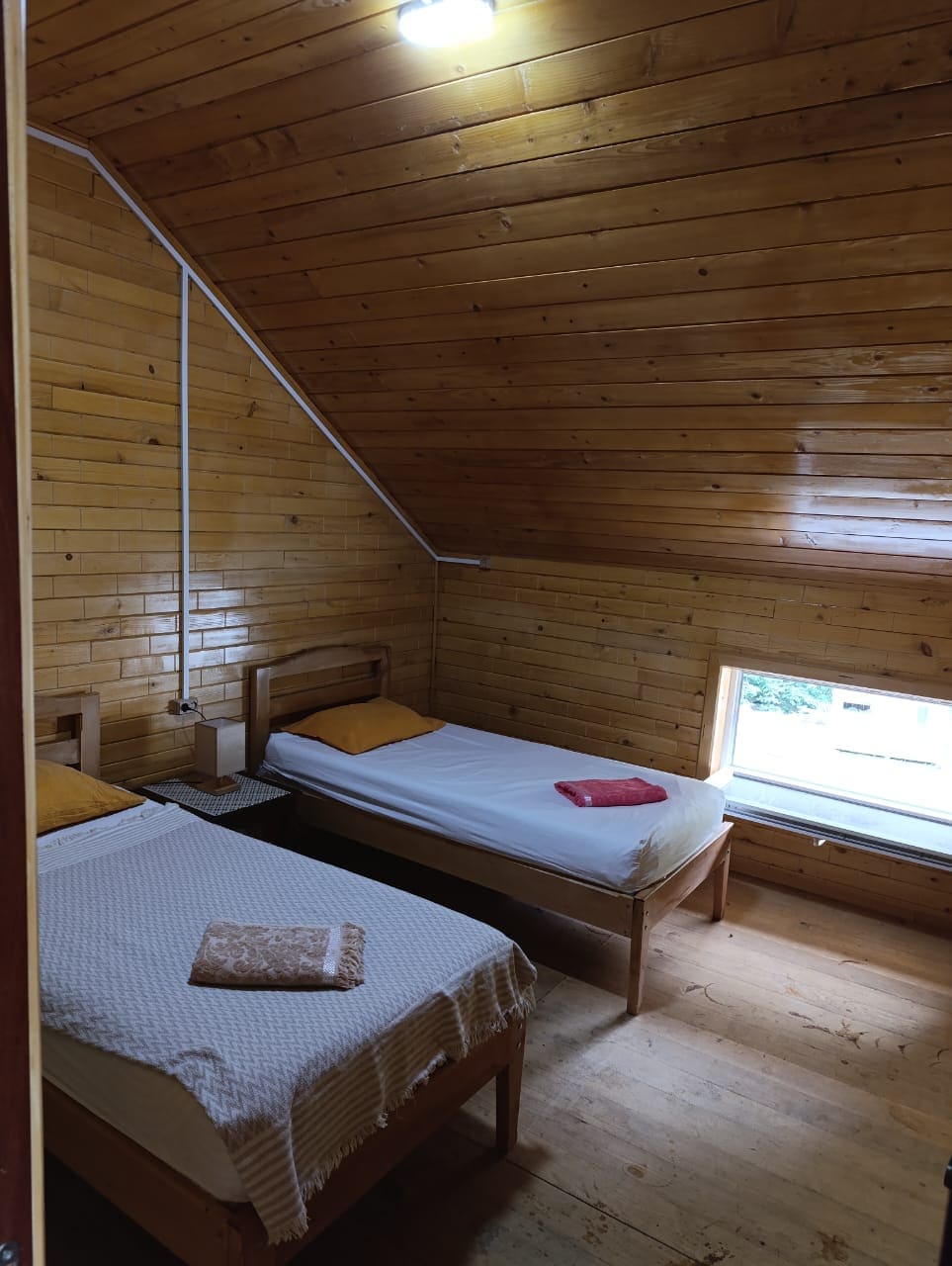 A cozy room features two single beds with simple linens. Wooden walls and ceilings contribute to a natural ambiance, while a window provides natural light. A small nightstand sits between the beds, adorned with a towel and a patterned cloth.
