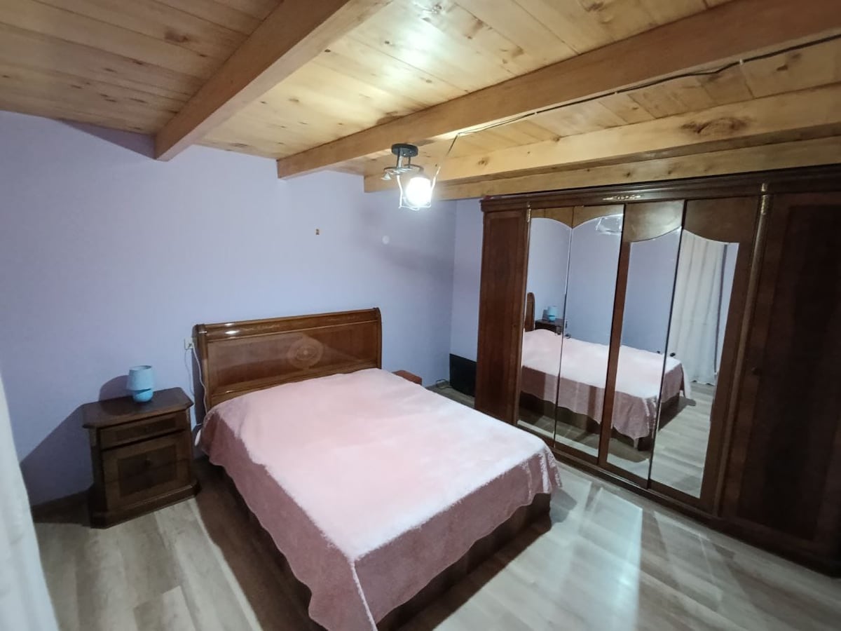 A sleeping area is furnished with a wooden bed covered by a light pink blanket. A wooden nightstand with a lamp stands beside the bed. A large mirrored wardrobe reflects the room's soft purple walls, with light wood beams visible on the ceiling.