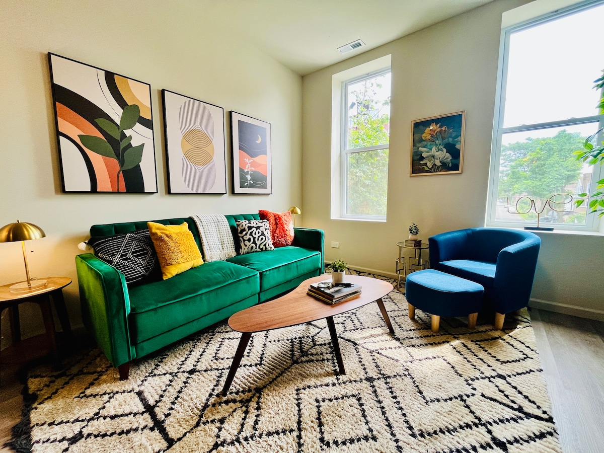 A welcoming living area is highlighted by a green sofa adorned with an assortment of colorful cushions. Above, modern art prints create visual interest on the walls, and a coffee table sits on a textured rug, complemented by natural light streaming through the large windows.