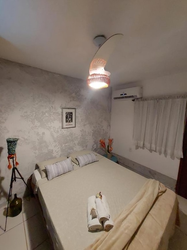 A cozy sleeping area features a spacious bed adorned with light linens and decorative towels. A stylish light fixture hangs from the ceiling, while a gentle air conditioning unit is mounted on the wall. Natural light filters through sheer curtains, enhancing the calm atmosphere.