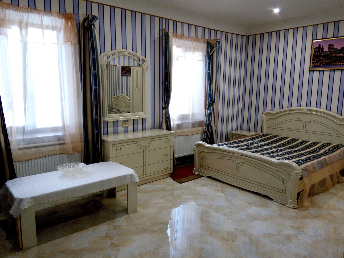 A room features a spacious bed with a patterned bedspread against striped wallpaper. Natural light is filtered through sheer curtains on two windows. A dressing table with a mirror and a small coffee table add functionality to the space. The glossy floor reflects the room's design.