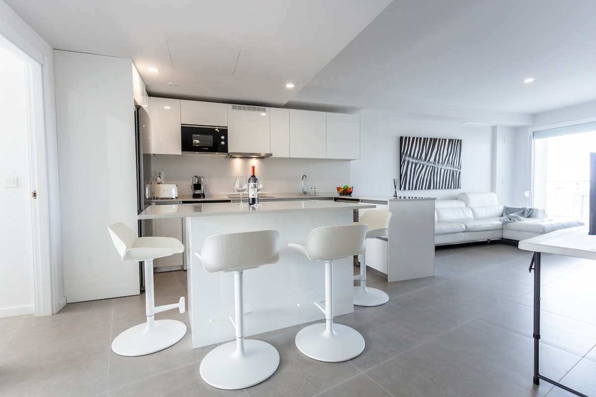 A modern kitchenette is highlighted by sleek white cabinetry and a central island with three high-backed stools. A set of appliances is visible, along with a dining area featuring a light-colored sofa. Natural light floods the space through large glass doors leading to a terrace.