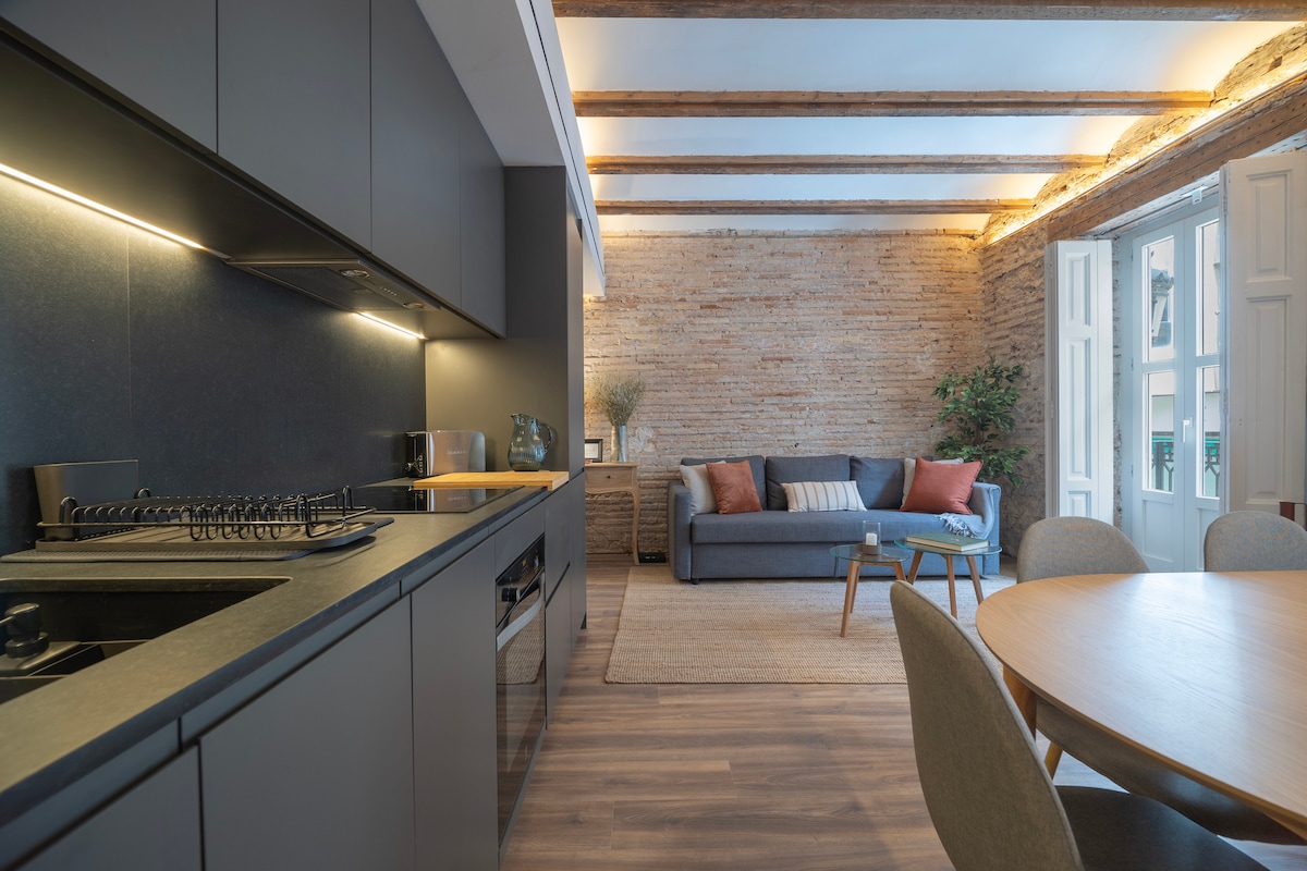 A modern kitchen with dark cabinetry and stainless steel appliances is complemented by a round wooden dining table. A cozy seating area with a grey sofa, adorned with colorful pillows, is illuminated by natural light from large open windows and highlighted by exposed wooden beams.