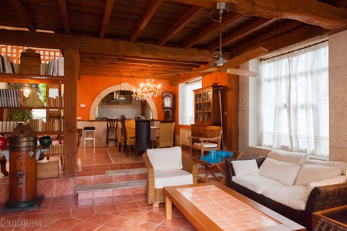 The spacious living area features a mix of natural materials and warm colors. A comfortable seating arrangement is present, accompanied by a coffee table. The area showcases a rustic kitchen in the background, illuminated by an elegant chandelier, with large windows allowing natural light.