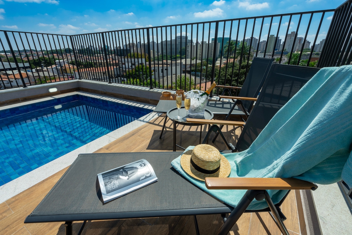 A sunlit terrace features a lounge chair with a light turquoise towel draped over it. A straw hat rests nearby, with a magazine placed on the ground. Two glasses and a small table accompany a sparkling pool, while a cityscape is visible in the background.