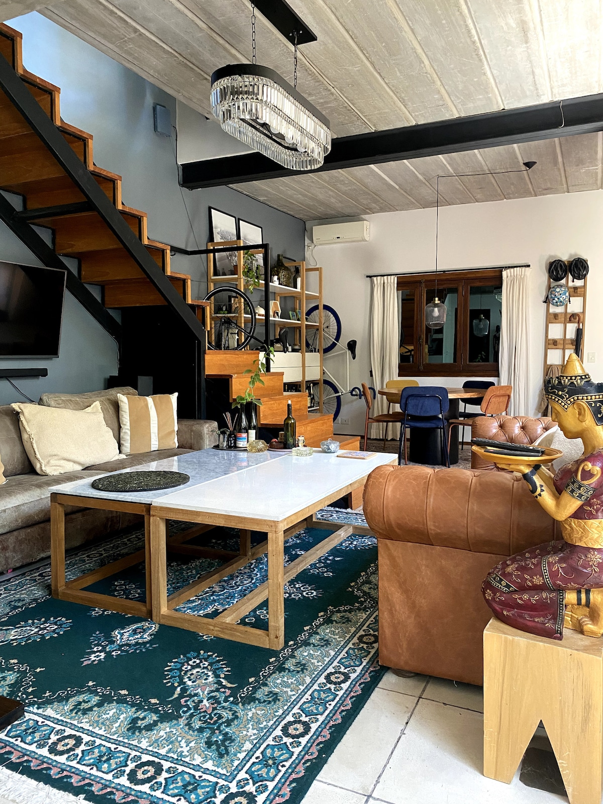 The living area features a modern coffee table set on a decorative rug, surrounded by a comfortable sofa and an armchair. A staircase leads to the upper floor, while a dining area is visible in the background. Large windows provide natural light, and a decorative statue enhances the space.