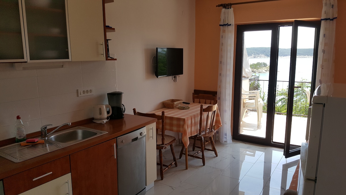 The kitchen and dining area feature wooden cabinetry and a tiled backsplash. A small table with checkered cloth is surrounded by four wooden chairs. A large window provides a view of the balcony, and a television is mounted on the wall.