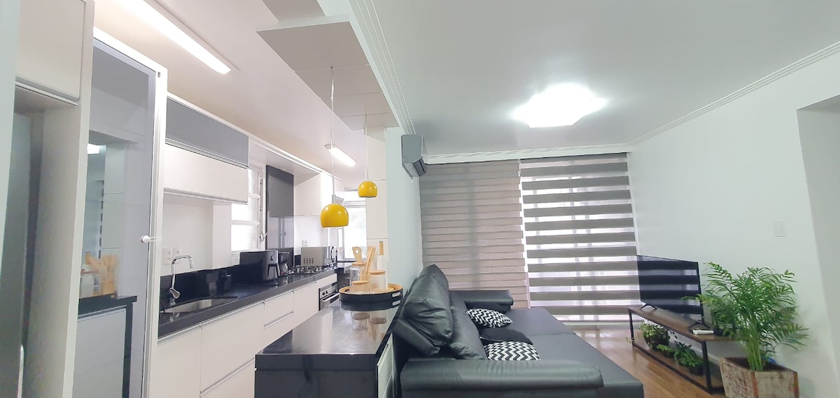 An open living area showcases a modern kitchen with sleek cabinetry and a dark countertop. A spacious black sofa is positioned in the foreground, accented by patterned cushions. A television sits across from the sofa, and large windows feature dual-tone blinds allowing natural light to filter through.