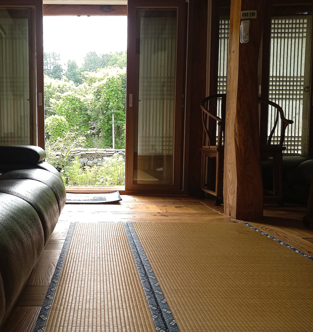 A serene interior space features a tatami mat floor that extends towards an entrance with sliding doors. Natural light filters in, illuminating a view of lush greenery outside. A dark sofa sits in the foreground, complemented by wooden chairs against the walls.