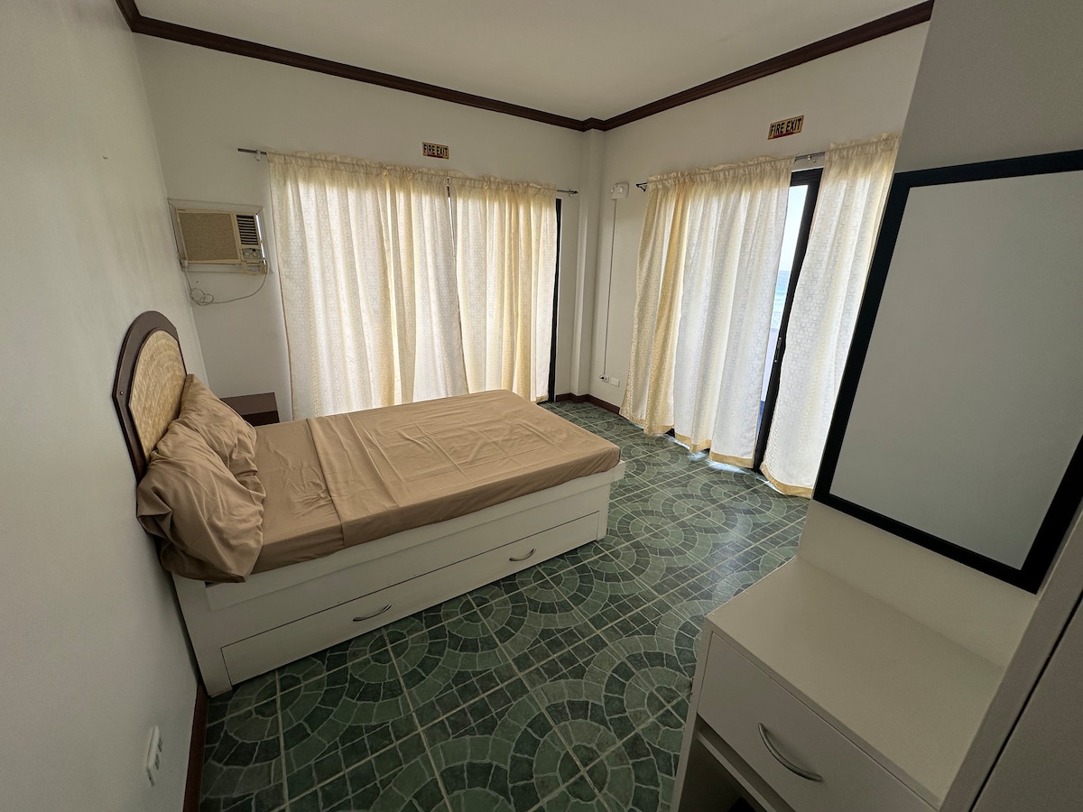 A simple bedroom features a single bed with a neutral-toned bedspread. Natural light filters through sheer curtains at two sliding glass doors, illuminating the tiled floor. A small dresser and a mirrored cabinet are positioned against the wall, enhancing the room's functionality.
