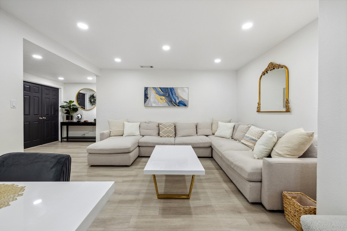 A spacious living area is showcased with a plush sectional sofa, accented by decorative pillows. A sleek white coffee table occupies the center, while soft lighting enhances the bright, neutral color palette. A decorative mirror and wall art add subtle interest to the room.