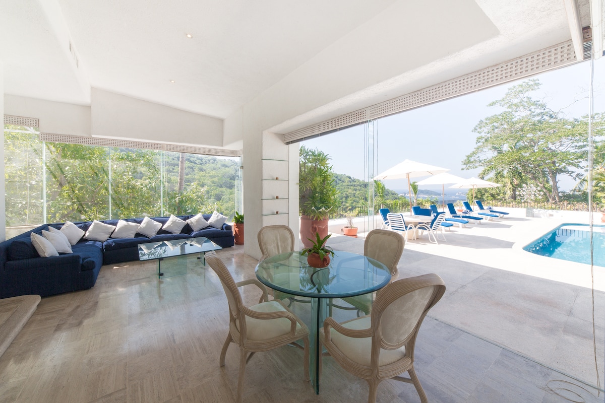 An open-concept living space is highlighted by a glass dining table surrounded by light-colored chairs. A comfortable blue sofa is positioned nearby, and large windows provide views of the swim area and lush greenery. Sun loungers and umbrellas are visible by the pool.