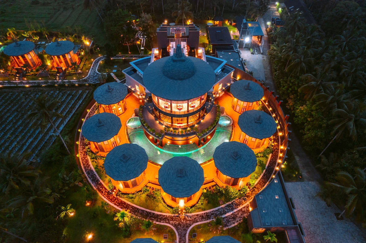 An aerial view of two-story villas displays a circular layout, highlighted by warm lighting. The structures are defined by their domed roofs and expansive windows, surrounded by lush greenery and pathways that weave through the landscape.