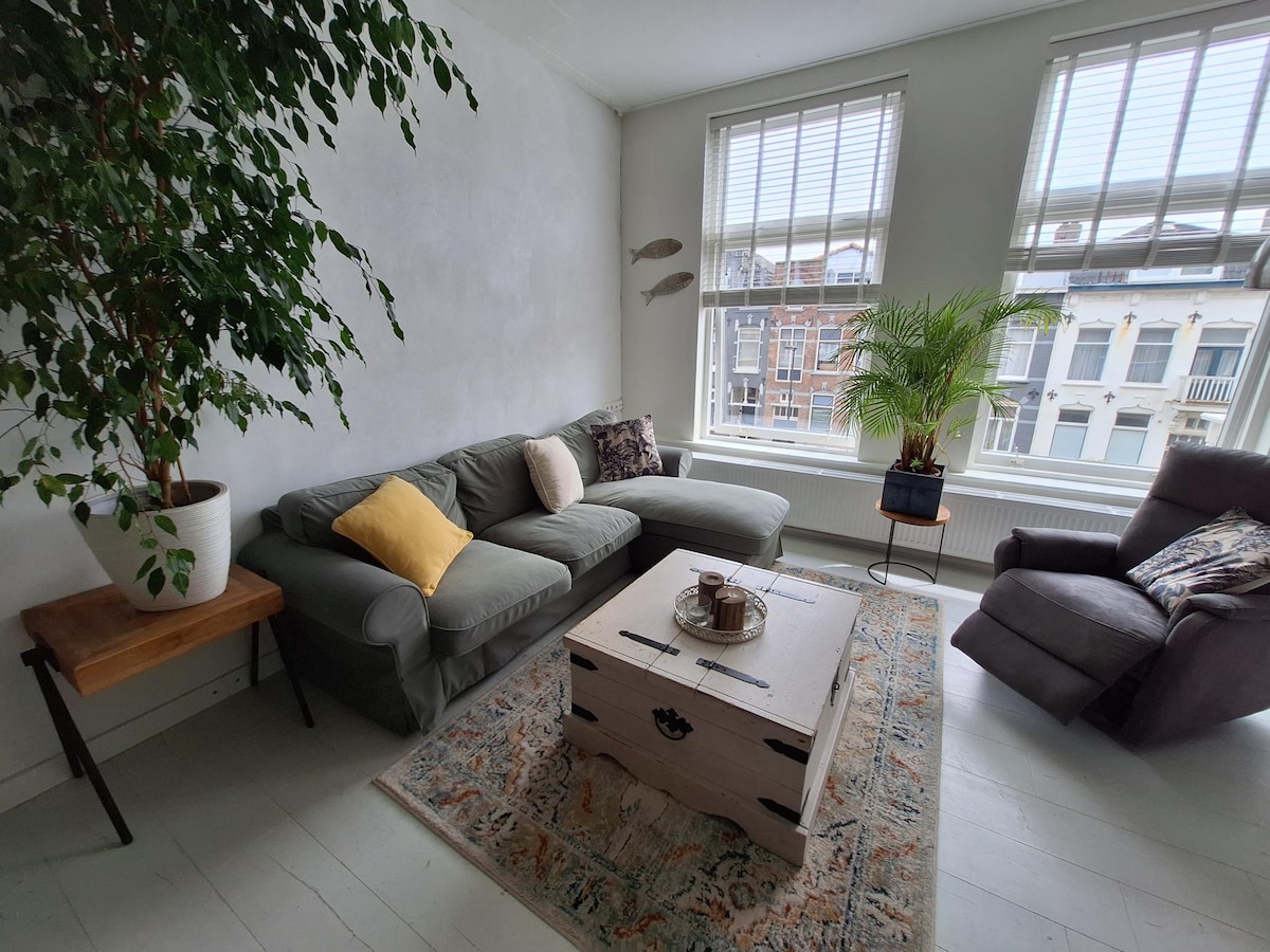 A comfortable seating area features a gray sectional sofa with an accent pillow, paired with a small wooden side table. A plant in a white pot adds a touch of greenery, while large windows allow natural light to fill the space. Decor is completed with a patterned rug.