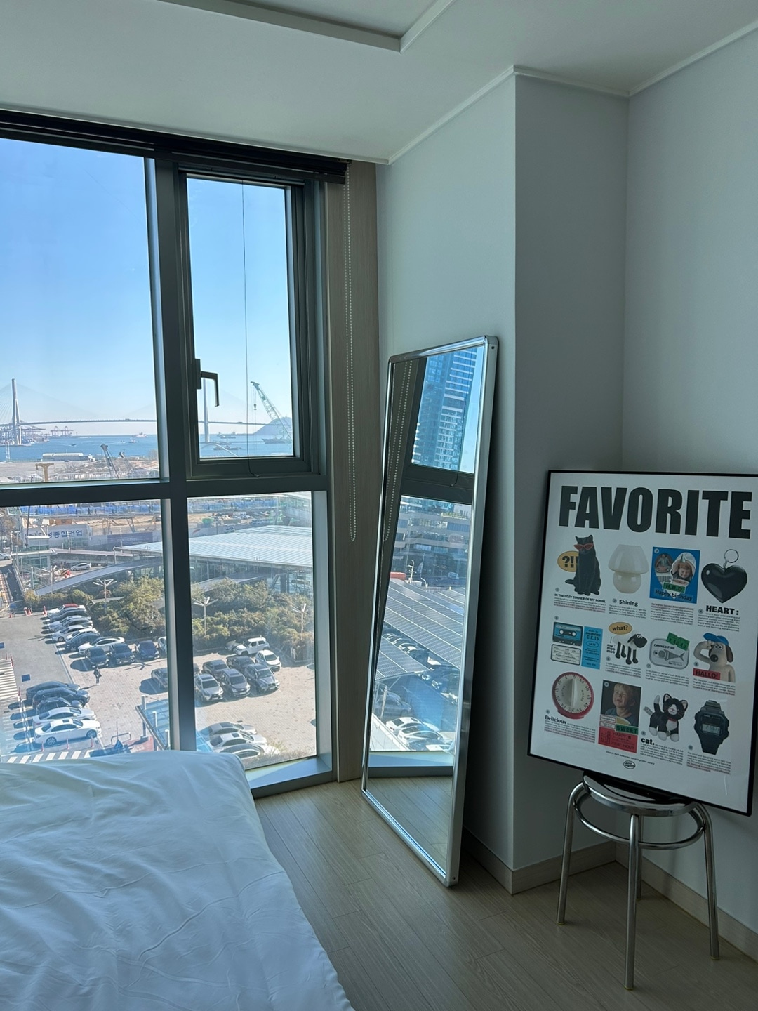 A bright room features large windows offering views of the city and waterfront. A full-length mirror stands beside a poster with various images and text. Soft bedding is arranged on a spacious bed, allowing for a relaxing atmosphere.