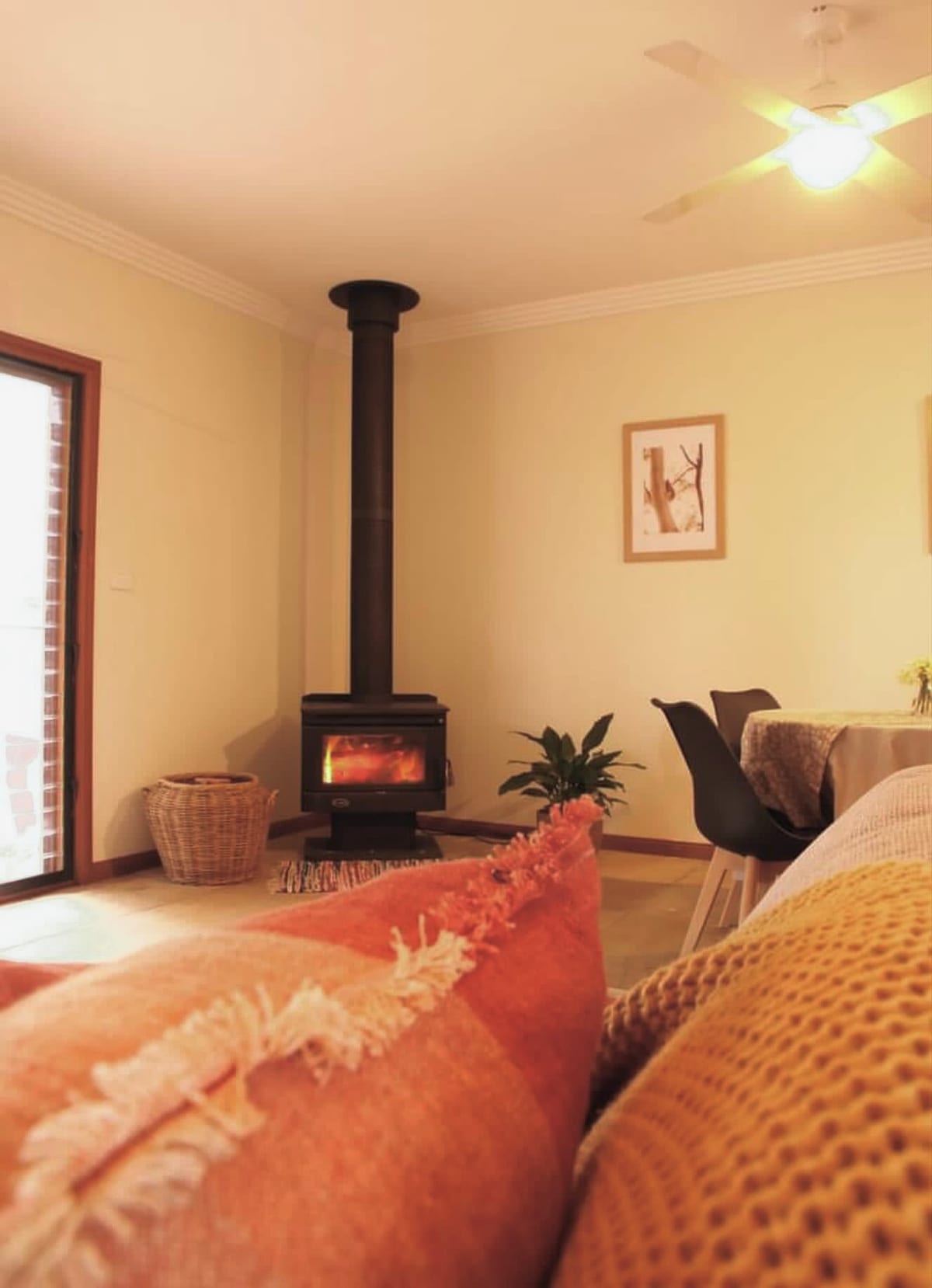 A cozy lounge area features a black wood stove providing warmth, surrounded by natural light from a nearby door. Soft, textured cushions in warm tones are placed on a comfortable couch, while a small indoor plant adds a touch of greenery. A dining table is visible in the background.