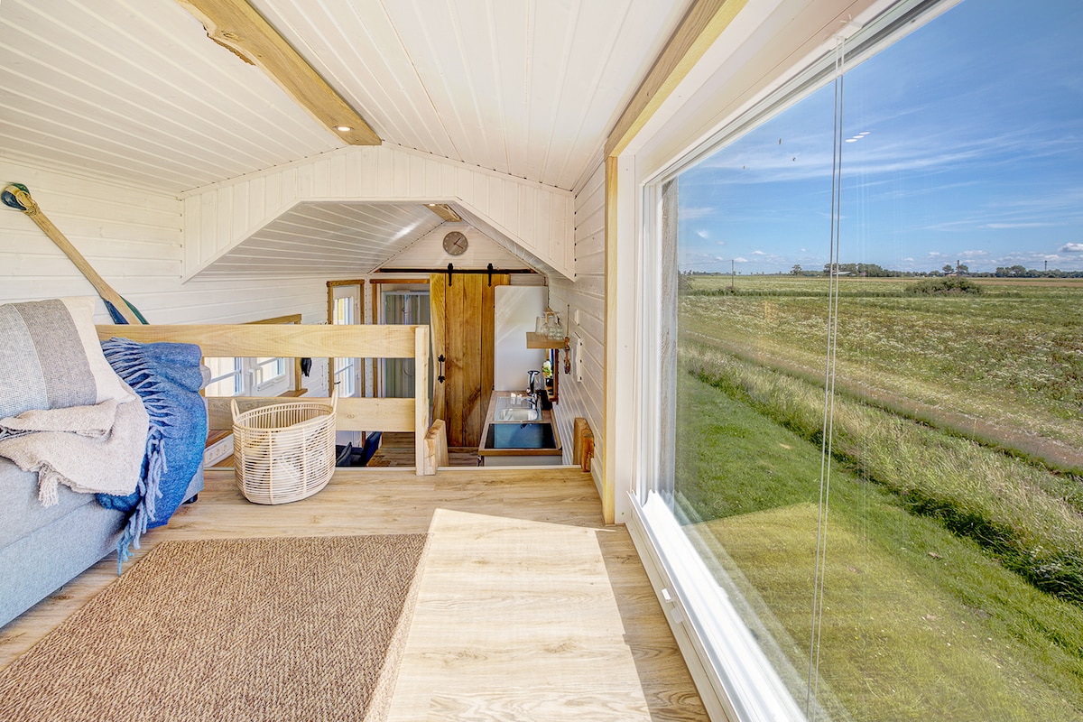 A cozy seating area is positioned near large windows, offering a view of the surrounding natural landscape. Soft light illuminates the wooden flooring and bright white walls. The atmosphere is complemented by a woven basket and a natural fiber rug, enhancing the space's rustic charm.