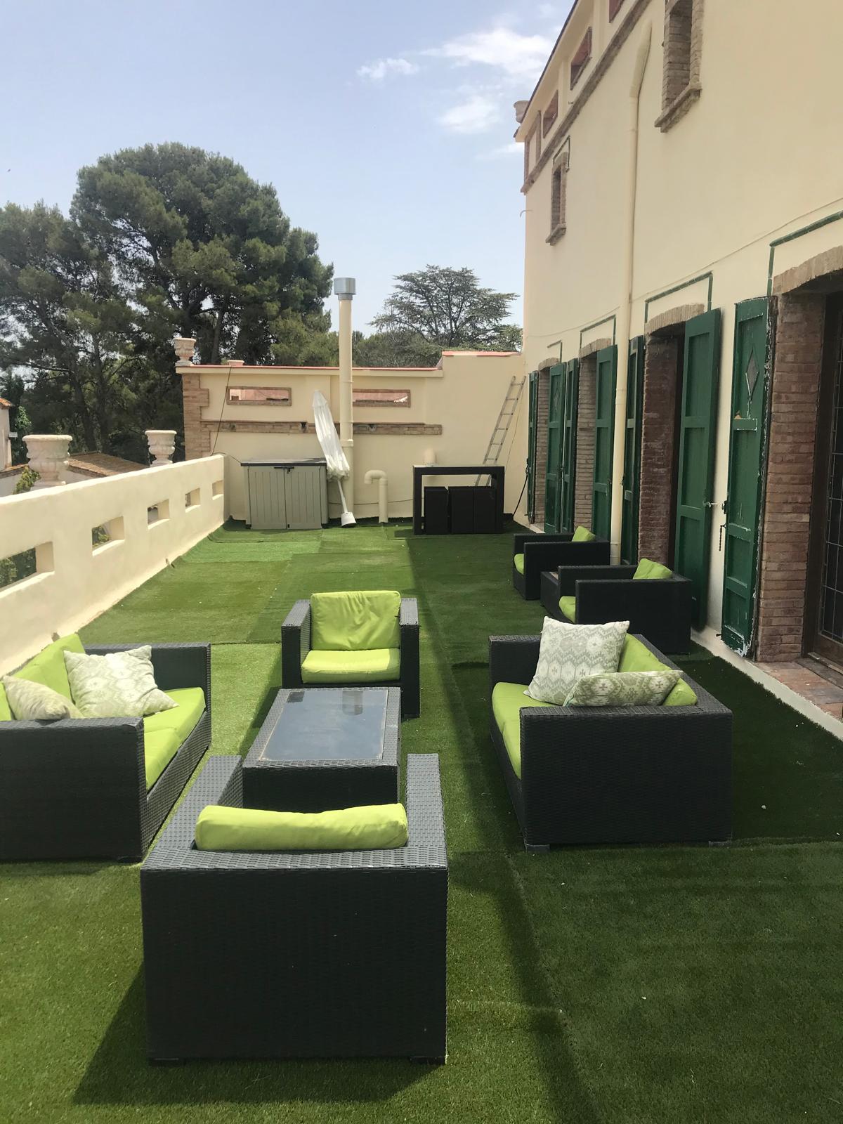 An outdoor terrace is displayed, featuring green artificial grass and multiple seating areas. Dark wicker seating with light green cushions surrounds a rectangular glass table. Large green shutters adorn the adjacent wall, while a ladder and storage unit are visible in the background.