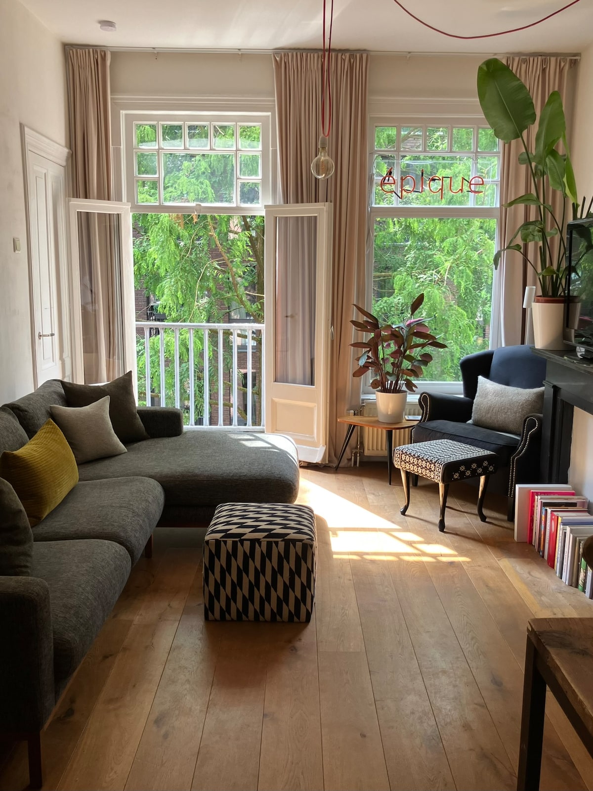 A spacious living room features large windows that allow natural light to fill the area. A comfortable grey couch is positioned invitingly, alongside a plush armchair. A patterned ottoman sits in front of the seating area, while a vibrant plant and bookshelves add warmth to the decor.