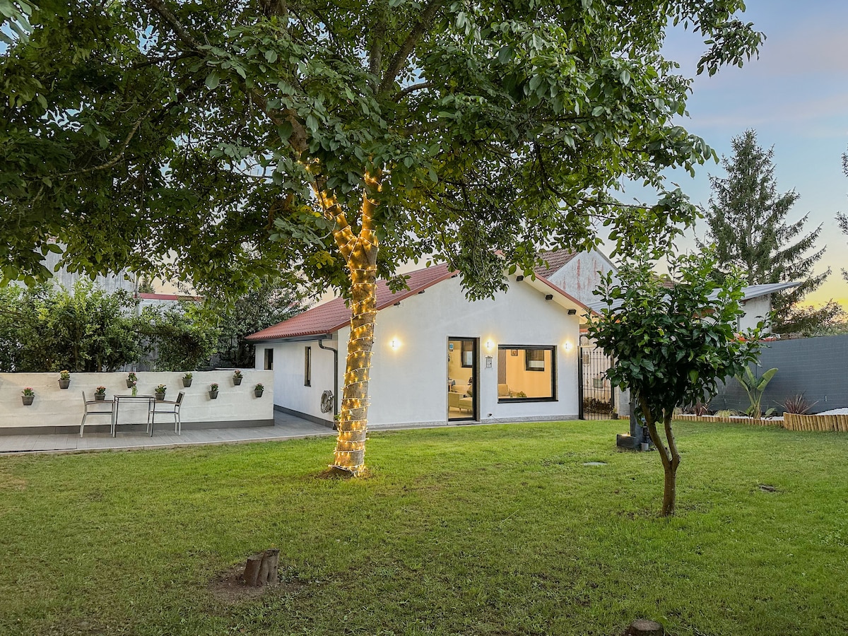A charming exterior view of the chalet features a well-lit white façade with dark trim, surrounded by lush green grass and trees. A cozy seating area is positioned under the tree, creating an inviting outdoor space for relaxation.