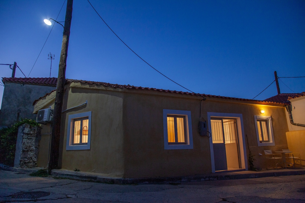 An exterior view of a charming, single-story building is captured at dusk, highlighting warm light emanating from the windows. The rustic yellow facade contrasts with the blue evening sky. A single wooden door is visible, alongside a small outdoor seating area with two chairs.