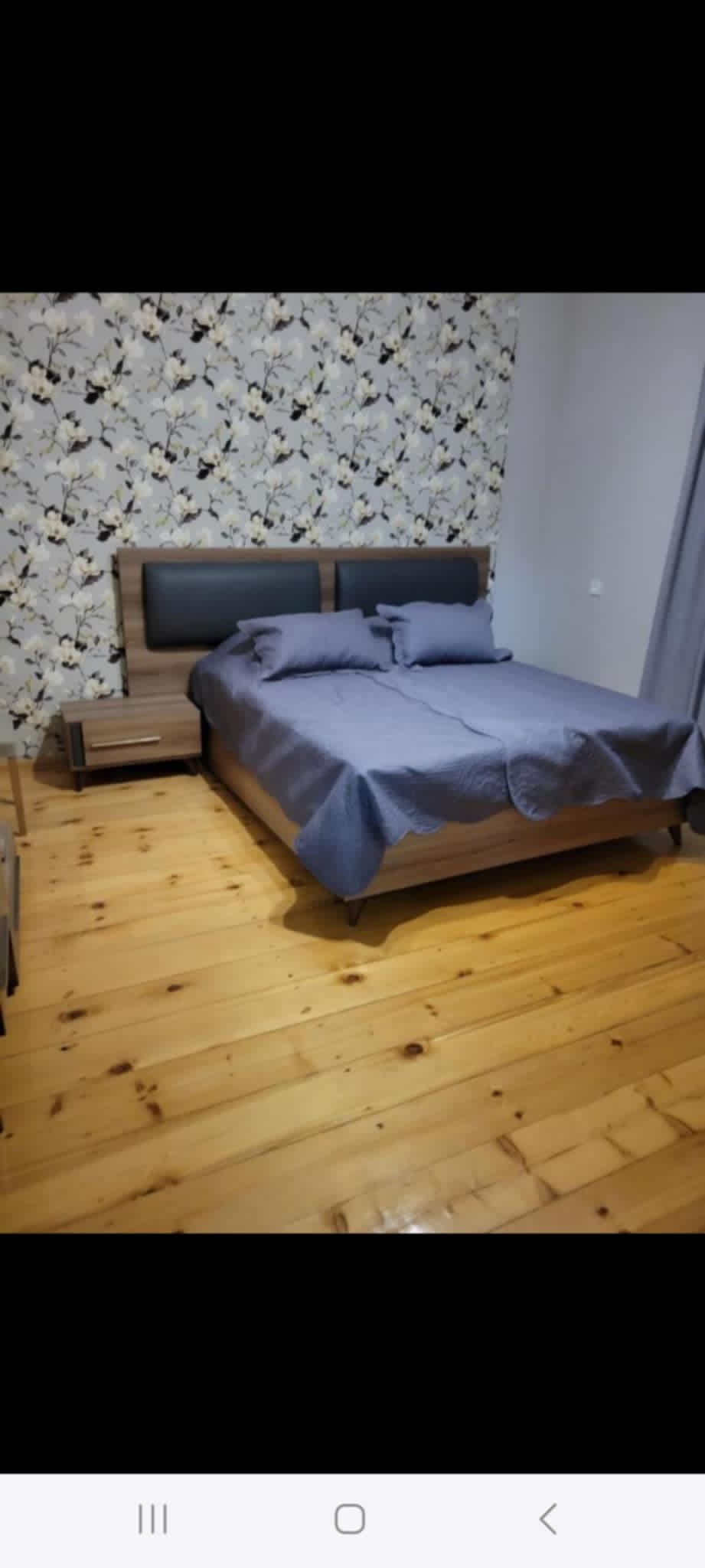 A spacious sleeping area features a bed with a dark grey coverlet and two matching pillows. A bedside table is present on one side, while a wooden floor enhances the room's warmth. A floral wallpaper adds a touch of detail to the background.