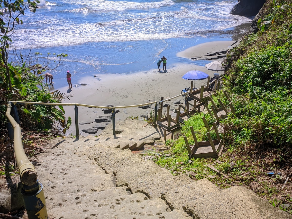 A set of stone steps leads down to a sandy beach, where gentle waves meet the shore. Several people are seen enjoying the beach activities, while beach chairs are positioned nearby under a large umbrella, surrounded by lush greenery and rocky formations.
