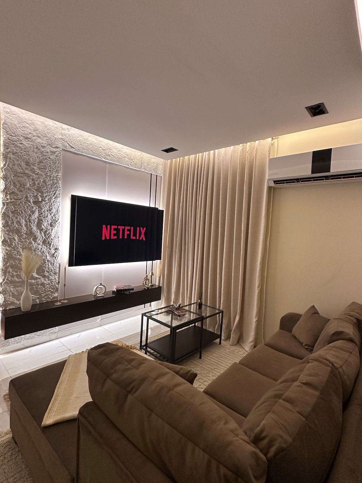 A comfortable seating area is highlighted by a plush sofa facing a modern television mounted on the wall. Soft lighting creates an inviting ambiance, while sheer curtains frame the space. A small coffee table sits in the center, completing the cozy arrangement.