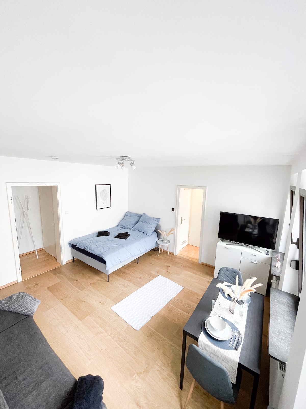 A bright living space features a blue daybed, a dark sofa, and a dining table set for four. Light wood flooring complements the neutral walls. A television is mounted on the wall, and a doorway offers access to additional areas of the apartment.