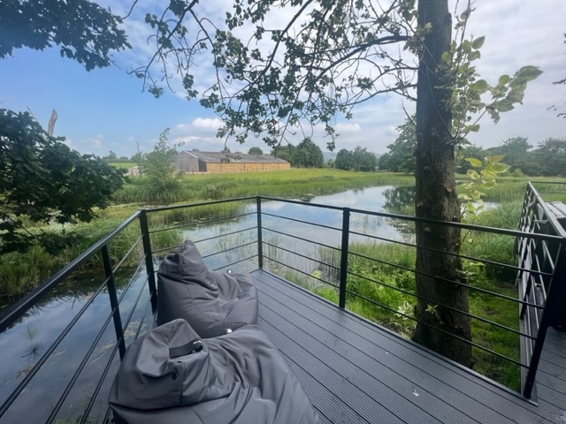 A spacious deck is featured, overlooking a natural pool surrounded by lush greenery. Comfortable bean bag seating is positioned for relaxation, with clear skies and gentle vegetation enhancing the serene ambiance of the countryside.