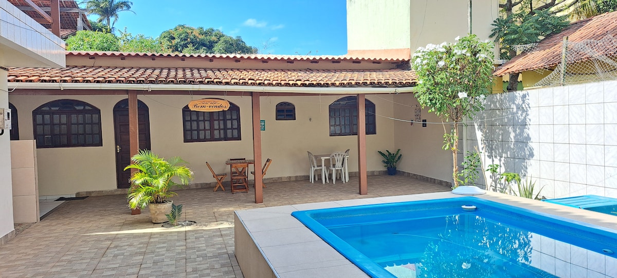 An outdoor area is shown, featuring a shared swimming pool bordered by tile. A covered patio with wooden posts includes a small table and chairs, surrounded by potted plants. Sunlight illuminates the space, highlighting the building's entrance with multiple windows.