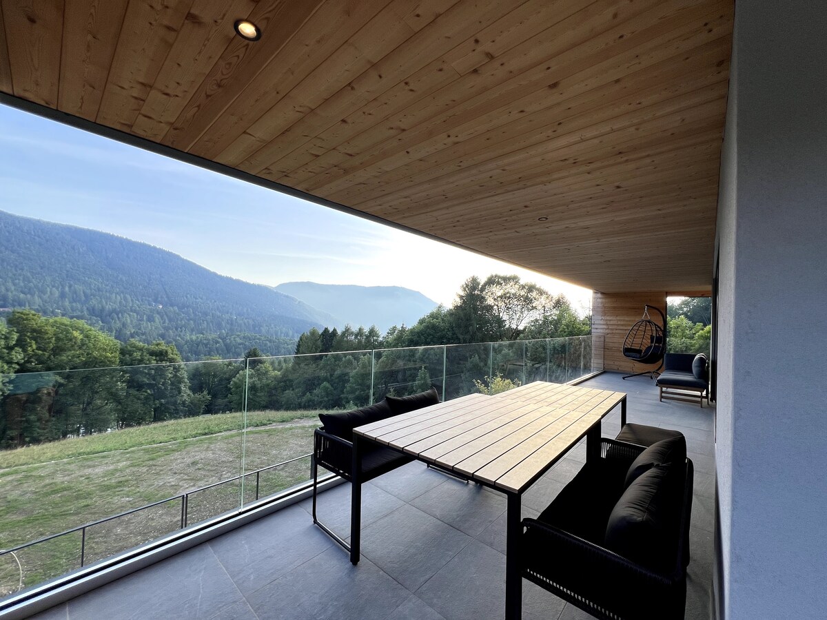 A spacious terrace features a wooden table alongside comfortable black seating, offering a panoramic view of the surrounding mountains and greenery. The ceiling is finished with natural wood, and a hanging chair adds a cozy element to the outdoor space.