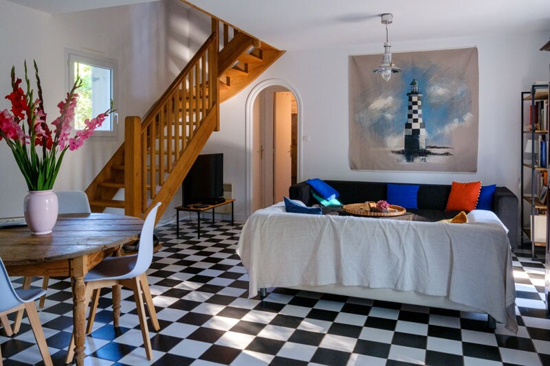 A bright living area features a checkerboard floor pattern in black and white. A wooden dining table and chairs sit beside a comfortable sofa adorned with colorful pillows. A large wall artwork of a lighthouse adds a coastal touch, while a staircase leads to the upper level.