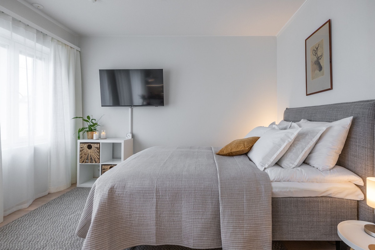 A cozy sleeping area is presented with a spacious bed adorned with white pillows and a light gray blanket. A flat-screen television is mounted on the wall, and a small side table with a lamp complements the room. Natural light filters through sheer curtains by the window.