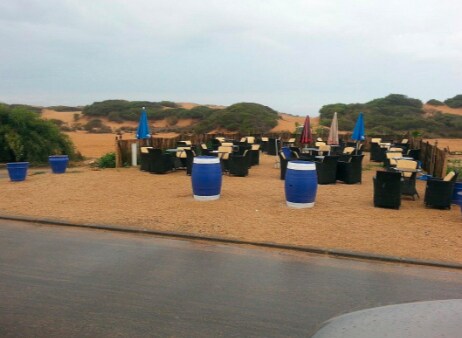A sandy outdoor seating area is visible, featuring multiple black chairs with light-colored tables. Blue barrels are positioned throughout, alongside colorful umbrellas in shades of blue, red, and beige. Distant dunes are seen in the background under an overcast sky.