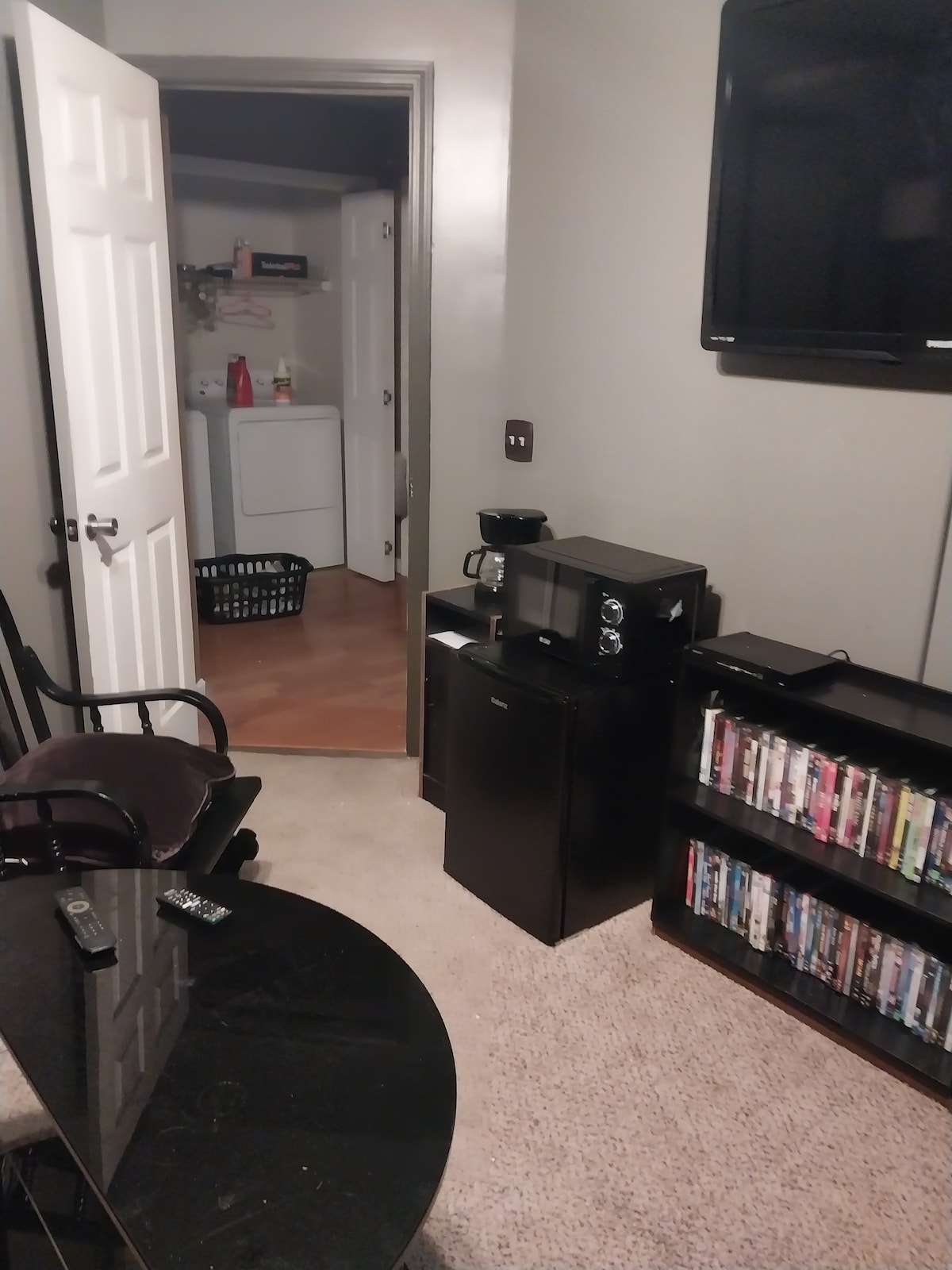 A cozy living area features a compact black coffee maker and a mini-fridge beside a sleek television. A collection of DVDs is neatly organized on a shelf. The doorway leads to a laundry area with a basket, adding functionality to the space.