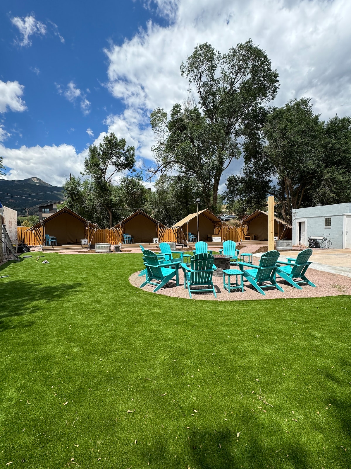 A sunlit outdoor space features vibrant blue seating arranged around a central fire pit on manicured green grass. Safari-style tents are situated in the background, surrounded by trees. The area is designed for relaxation and social interaction, complemented by a welcoming atmosphere.