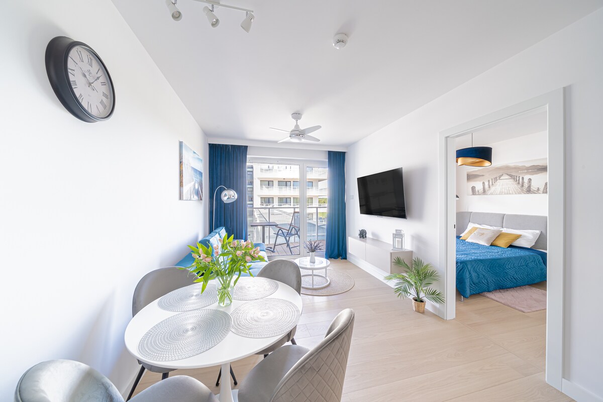 The living area displays a round dining table surrounded by four chairs, beside a ceiling fan and wall clock. A large window offers access to a balcony, while a cozy sleeping area is visible in the background, featuring a bed dressed in serene tones.
