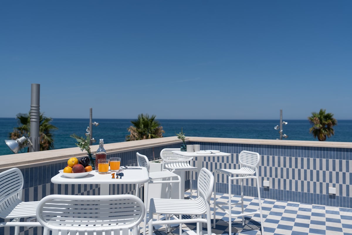 A shared terrace offers stunning sea views, featuring two tables surrounded by white chairs. A tray with fruits and beverages is seen on one table, while sunlight illuminates the tiled flooring, enhancing the outdoor setting.